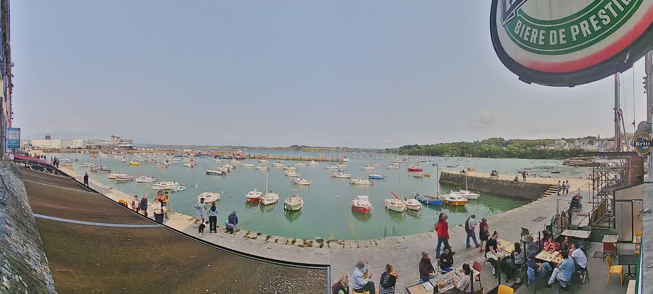 Many small boats are moored in a calm harbor on a sunny day, with people gathered on a nearby quayside at an outdoor café.