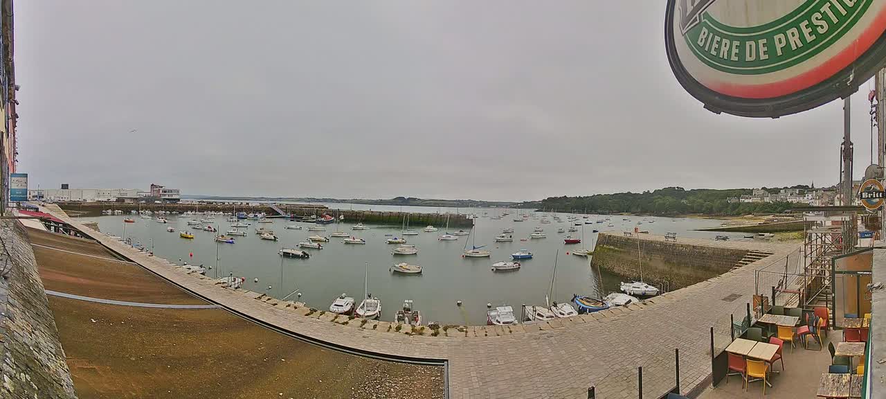 A harbor filled with numerous small boats on a cloudy day, viewed from an elevated position overlooking a stone paved area with outdoor seating.