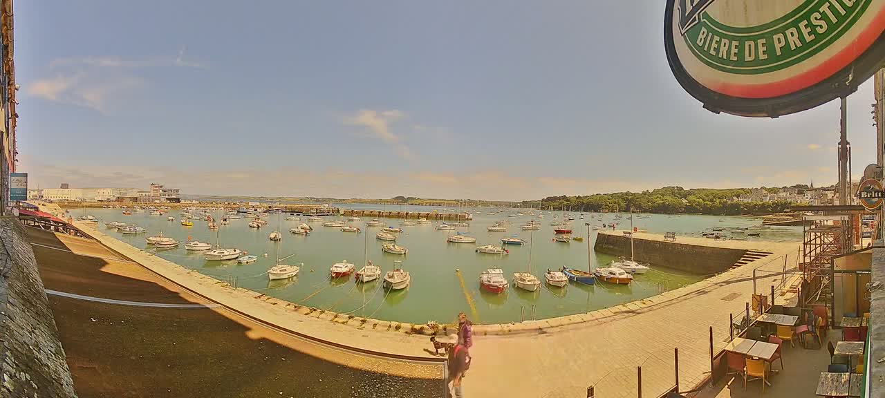 A sunny day shows a harbor filled with numerous small boats, a stone walkway, and a few people walking along the waterfront near outdoor seating.
