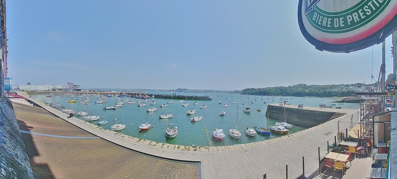 A harbor filled with numerous sailboats on a sunny day, viewed from an elevated position overlooking a stone quay and outdoor seating area.