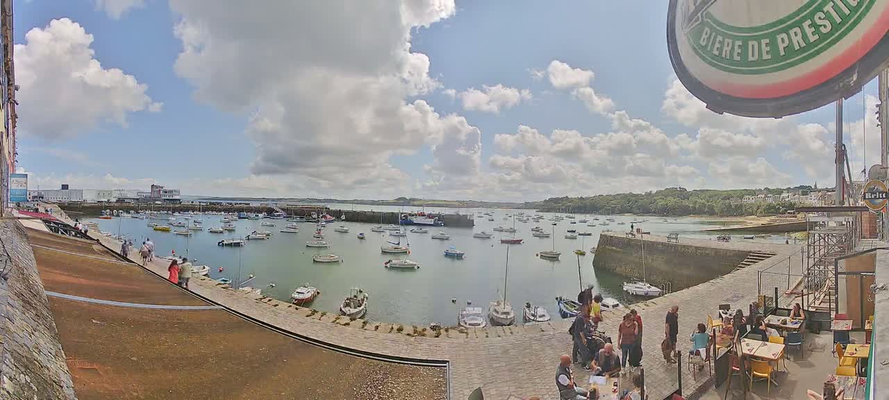 A harbor filled with numerous boats under a partly cloudy sky, with people gathered near a stone quayside and outdoor seating area.