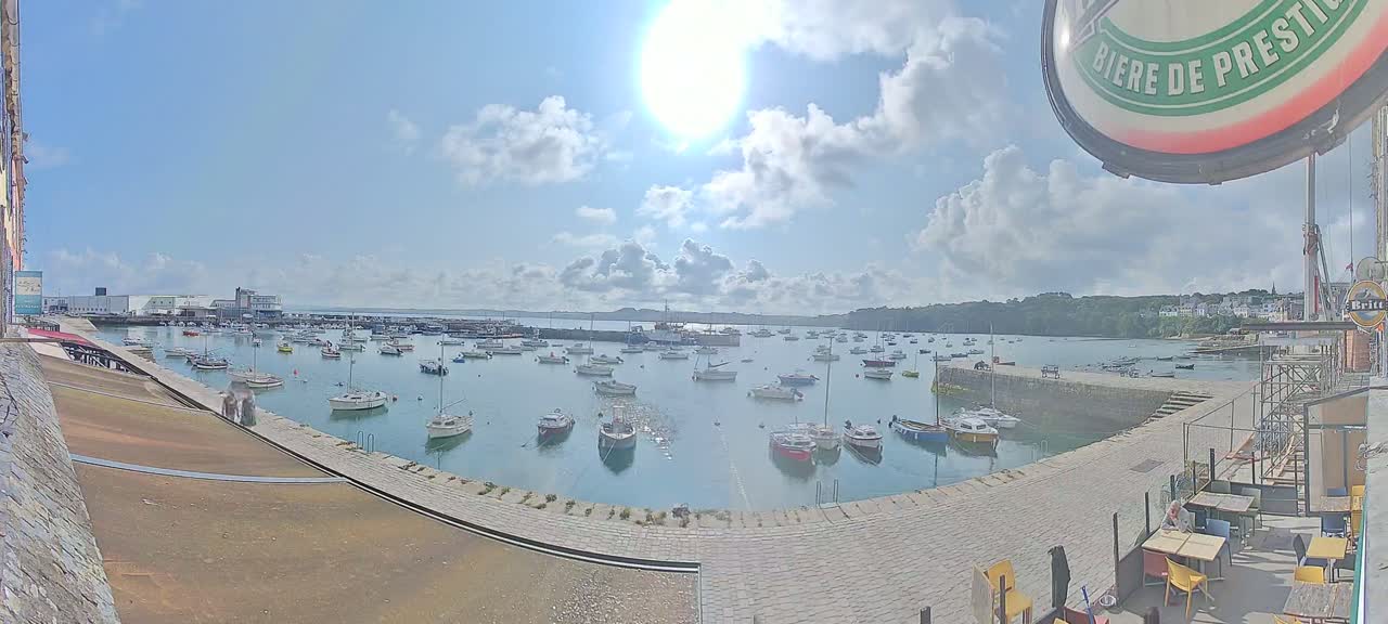 A sunny day with partly cloudy skies shows a harbor filled with numerous small boats, and a stone quay with outdoor seating areas.