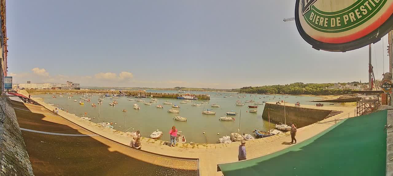A sunny day at a harbor filled with numerous boats, with people standing on a stone wall overlooking the water.