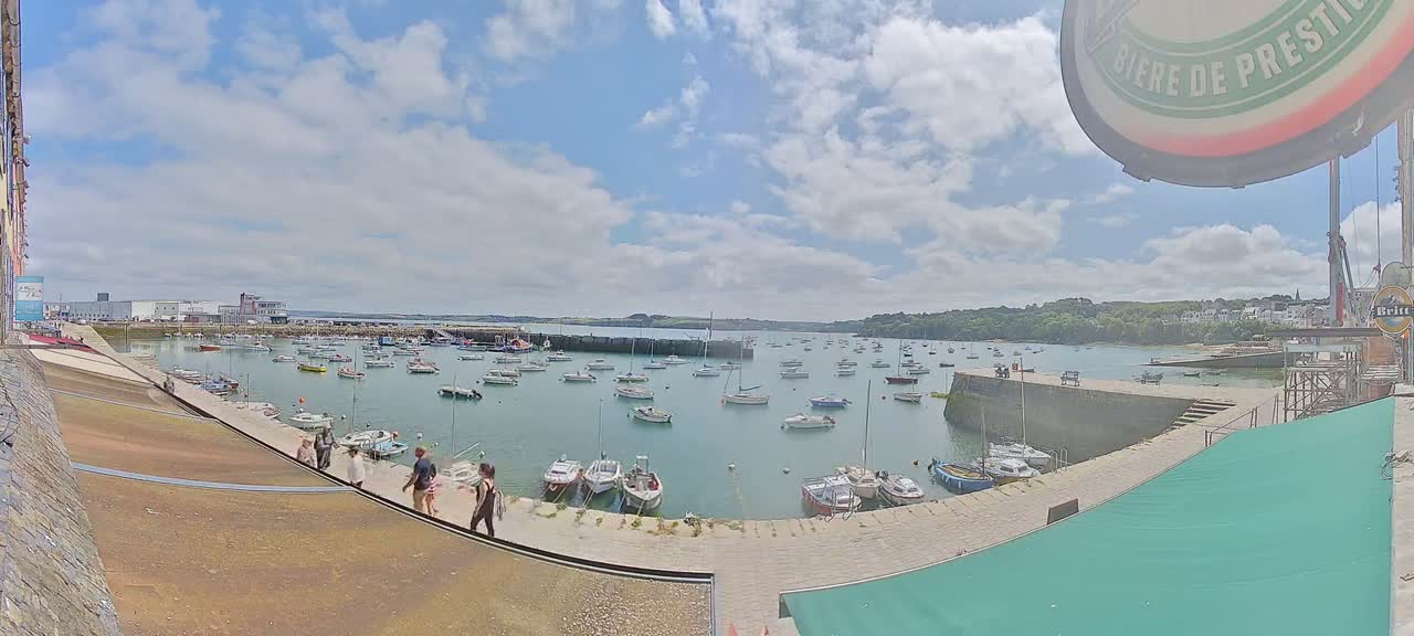 A harbor filled with numerous boats is viewed from a slightly elevated position on a partly sunny day with some fluffy clouds.