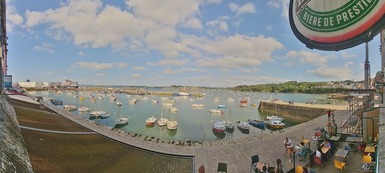 A harbor filled with numerous boats is seen under a partly cloudy sky, with people sitting at outdoor tables in the foreground.
