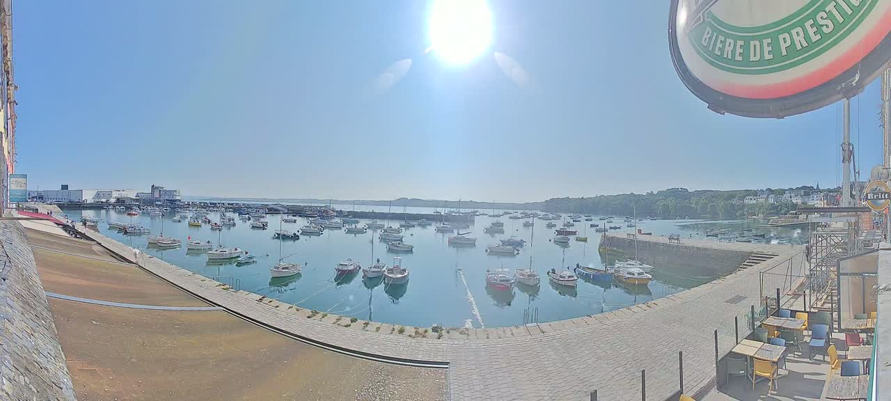 A sunny harbor filled with numerous small boats, a stone walkway, and outdoor seating.