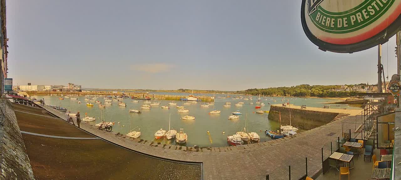 A sunny harbor filled with numerous small boats is viewed from a high vantage point, overlooking a paved area with outdoor seating.