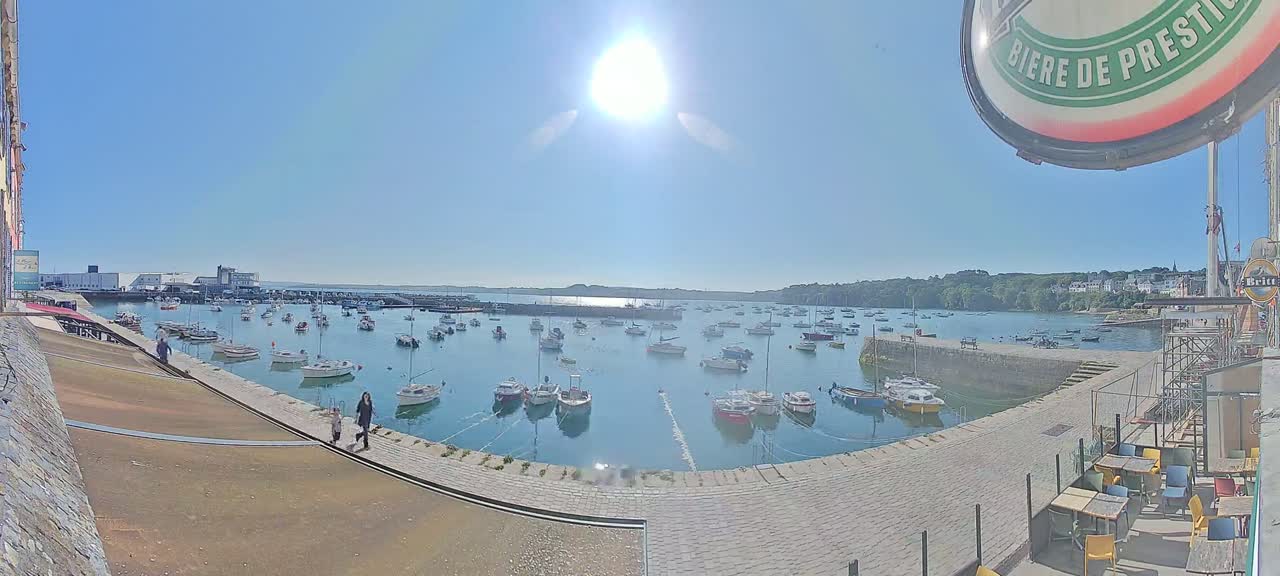 A sunny day at a harbor filled with numerous boats, overlooked by a stone walkway and outdoor seating area.