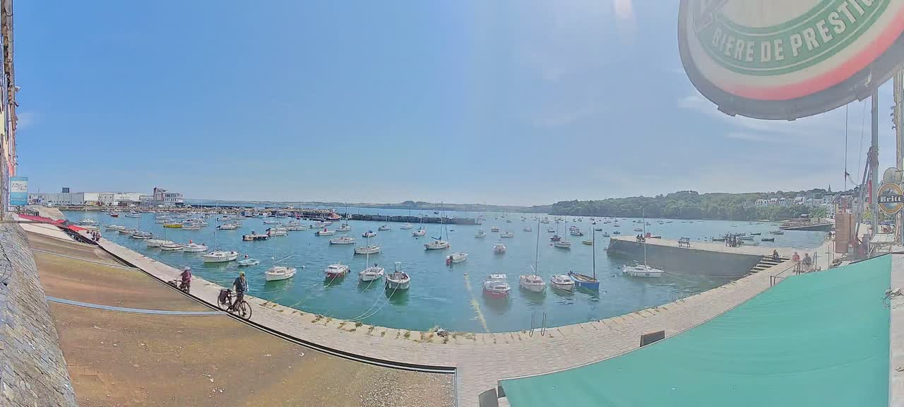 A sunny harbor filled with numerous sailboats and motorboats, with people walking and cycling along a paved waterfront.