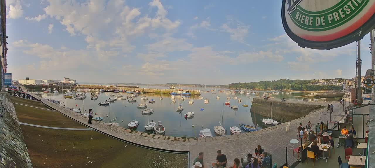 A harbor filled with numerous boats under a partly cloudy sky, with people relaxing at outdoor seating near the water's edge.