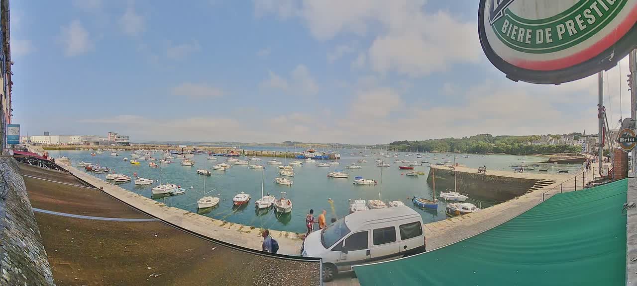 A sunny day reveals a harbor filled with numerous small boats, a van parked nearby, and several people standing on a stone quay overlooking the water.