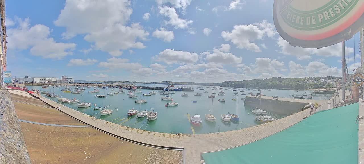 A sunny day with partly cloudy skies shows a harbor filled with numerous small boats and yachts, surrounded by buildings and green hills.