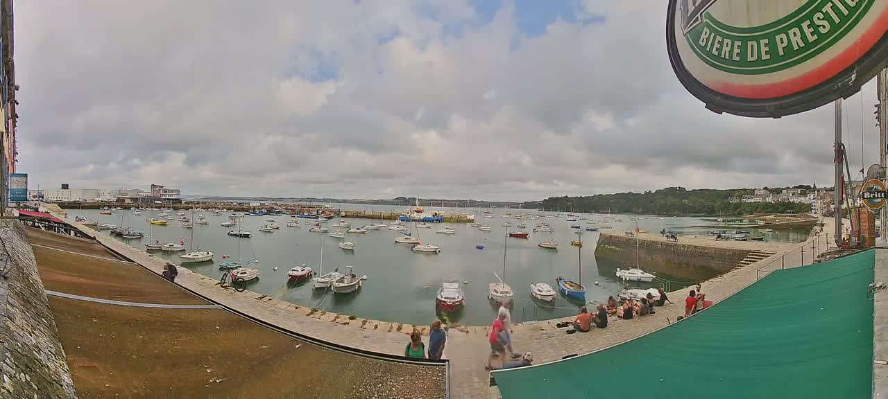 A harbor filled with numerous boats is viewed from a high vantage point on an overcast day, with people relaxing on the harbor wall.