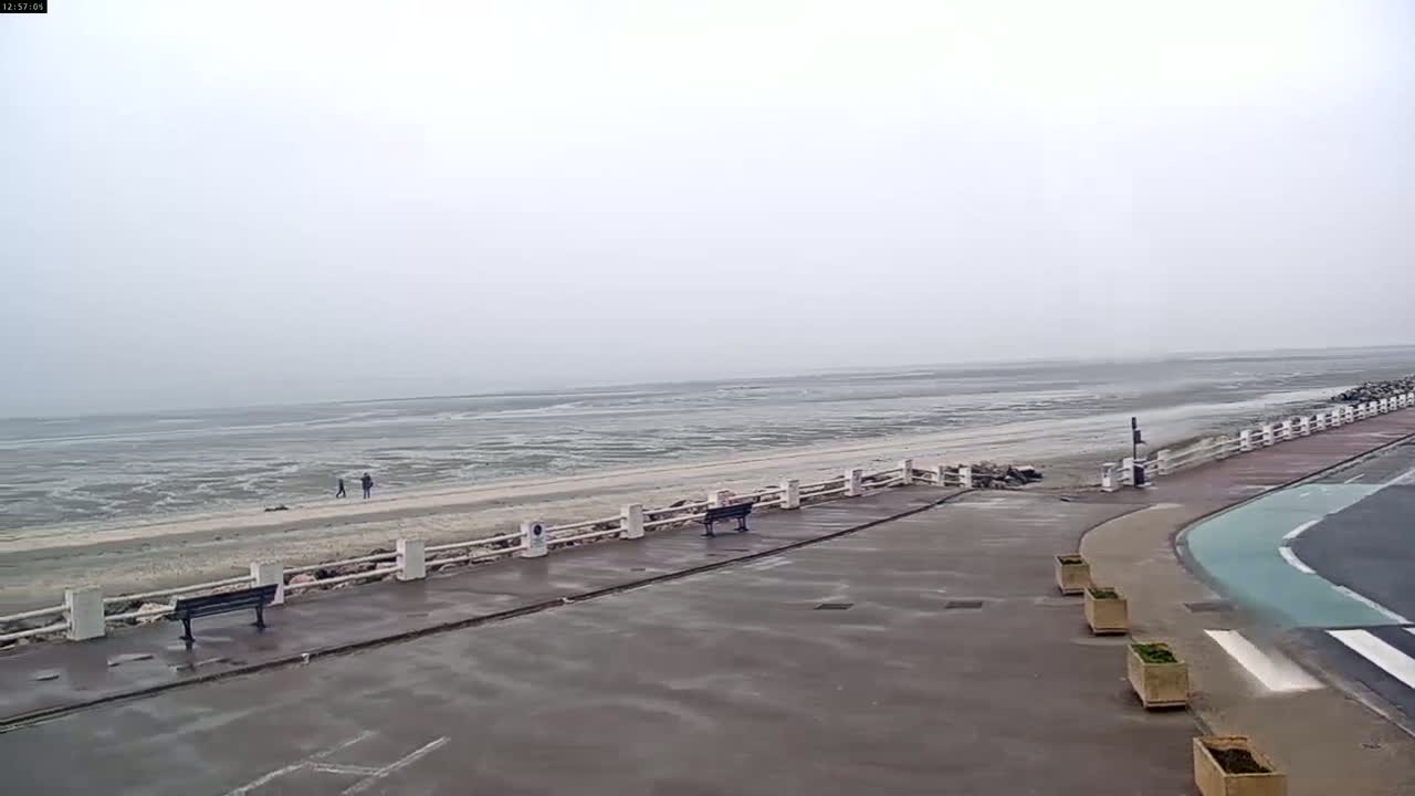 A mostly overcast day shows a wide, flat expanse of beach and ocean, with a paved walkway featuring benches and planters lining the ocean side.