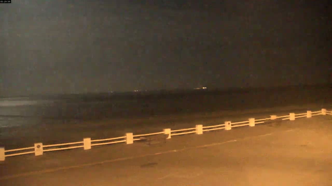 A dark, nighttime beach scene shows a low, double-railed fence separating a road from the dark water.