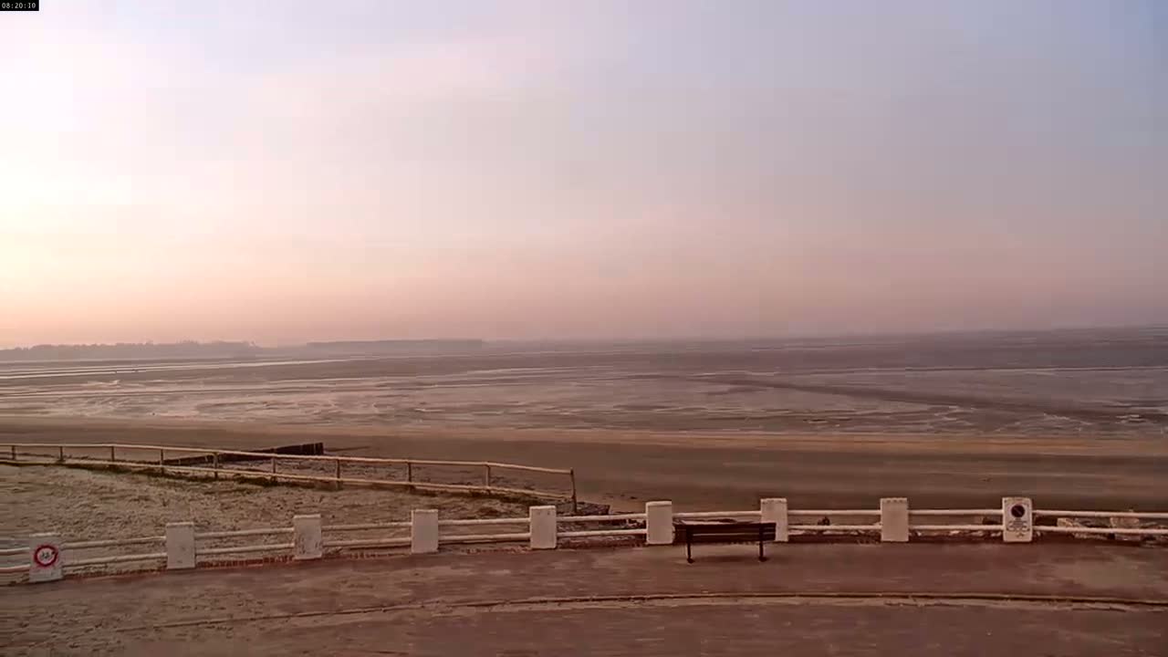 A hazy, pinkish sunrise illuminates a wide expanse of mudflats stretching to a distant shoreline, viewed from behind a low white fence and a park bench.