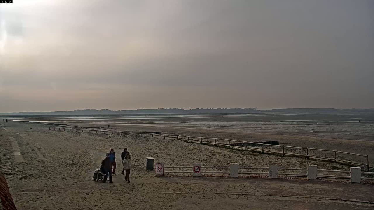 A sandy beach stretches out to a wide expanse of mudflats under a cloudy sky, with a few people walking near a low fence.
