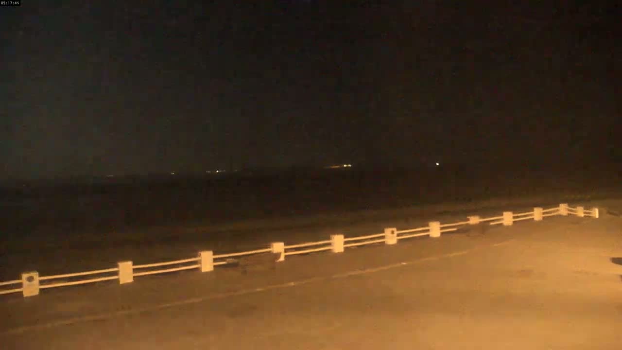 A nighttime view of a dark body of water, possibly the ocean, stretches to a distant horizon with faint lights visible, overlooked by a low, white railing along a roadway.
