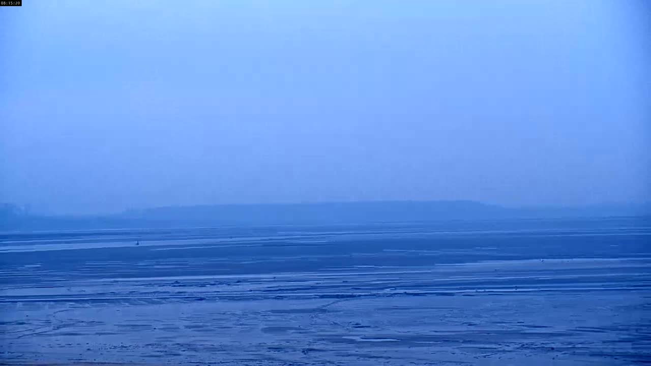 A hazy, blue sky overlooks a vast, flat expanse of dark blue water that appears to be receding, revealing a textured, muddy shoreline.