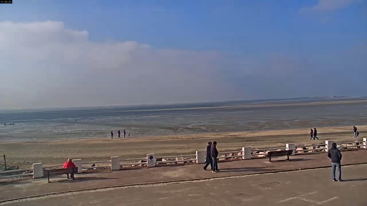 A mostly sunny day at a beach shows a wide expanse of exposed sand and shallow water, with several people walking on the sand and along a paved walkway near a low white fence.