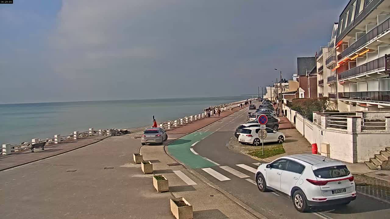 A seaside road with parked cars and pedestrians, under a partly cloudy sky, next to a calm ocean.