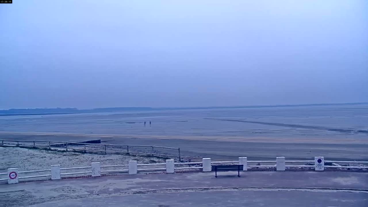 A wide, flat expanse of pale mudflats stretches to a hazy horizon under a muted, overcast sky, with a low white fence and bench in the foreground.