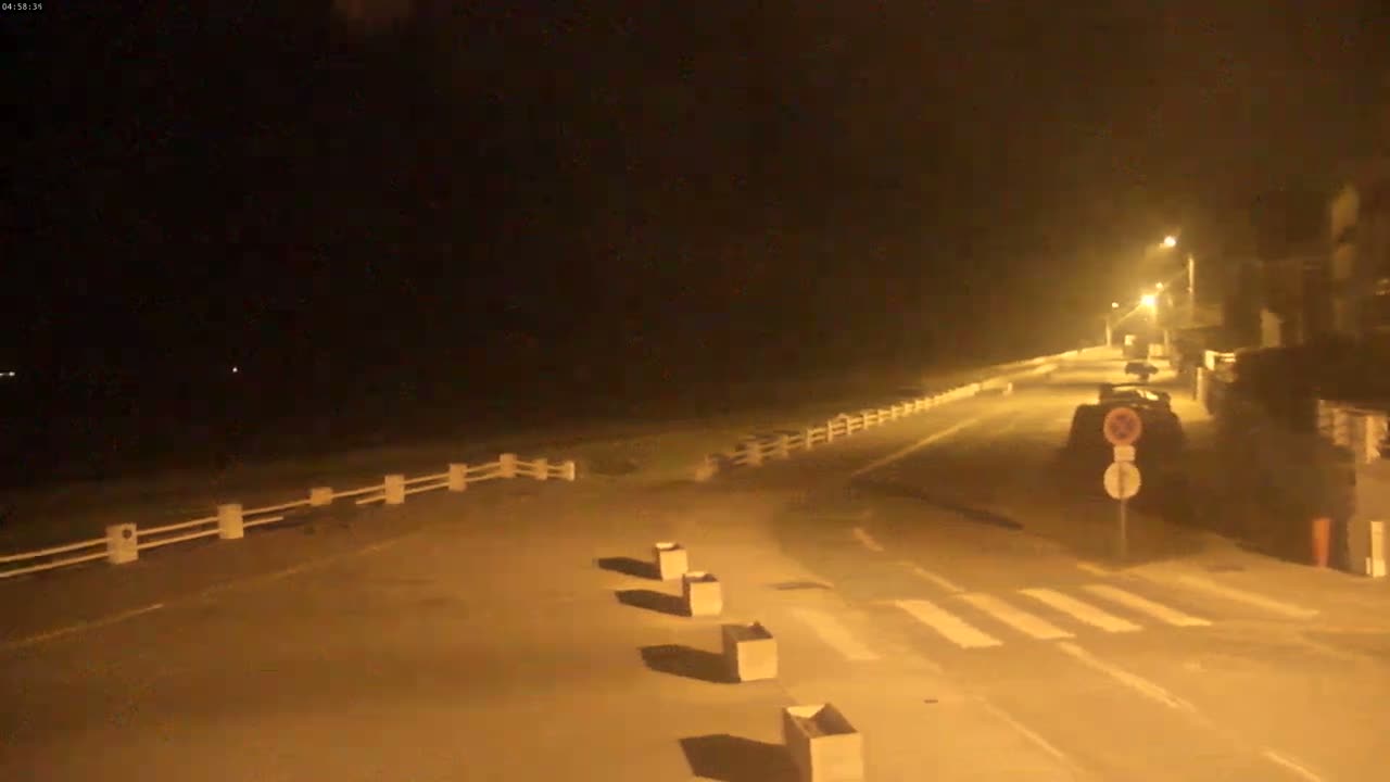 A nighttime, dimly lit road with a low white fence alongside it, leading toward buildings under a dark sky.