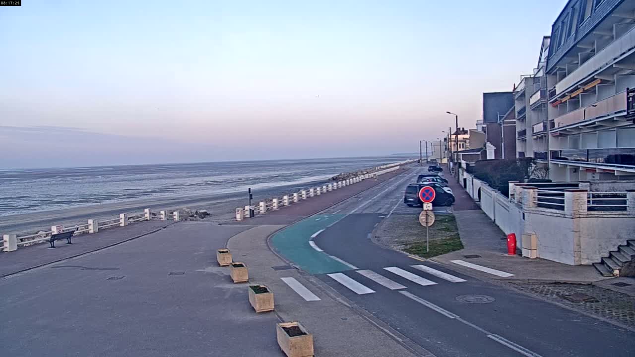 A coastal road with a bike lane curves toward a beach with a low tide under a pale, clear sky.