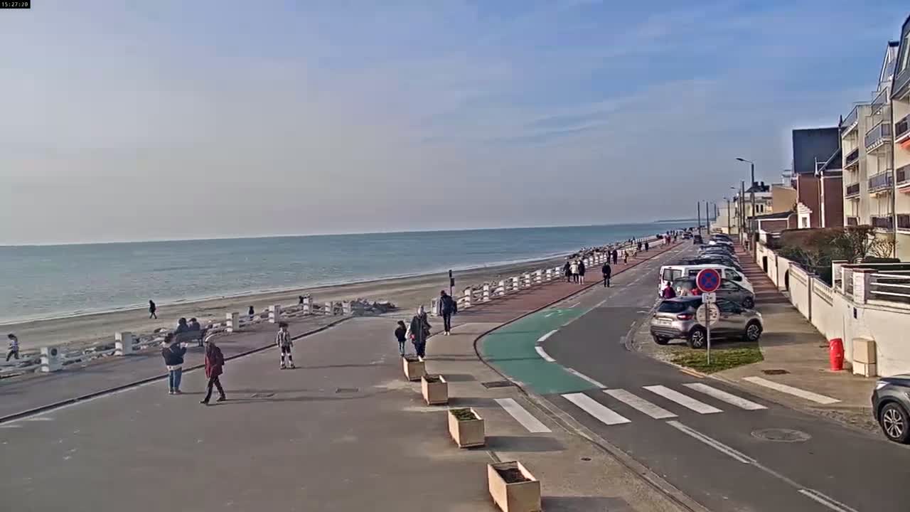 A paved walkway and road run alongside a sandy beach and calm ocean, under a mostly clear sky, with several pedestrians and parked cars visible.