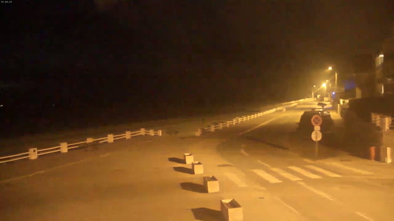 A nighttime street scene shows a sparsely-trafficked road with a white fence bordering a dark expanse, possibly the ocean, under hazy or foggy conditions.