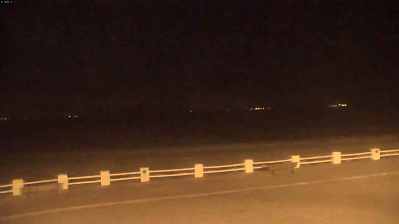 A nighttime view of a road with a low white fence, overlooking a dark body of water with distant lights.