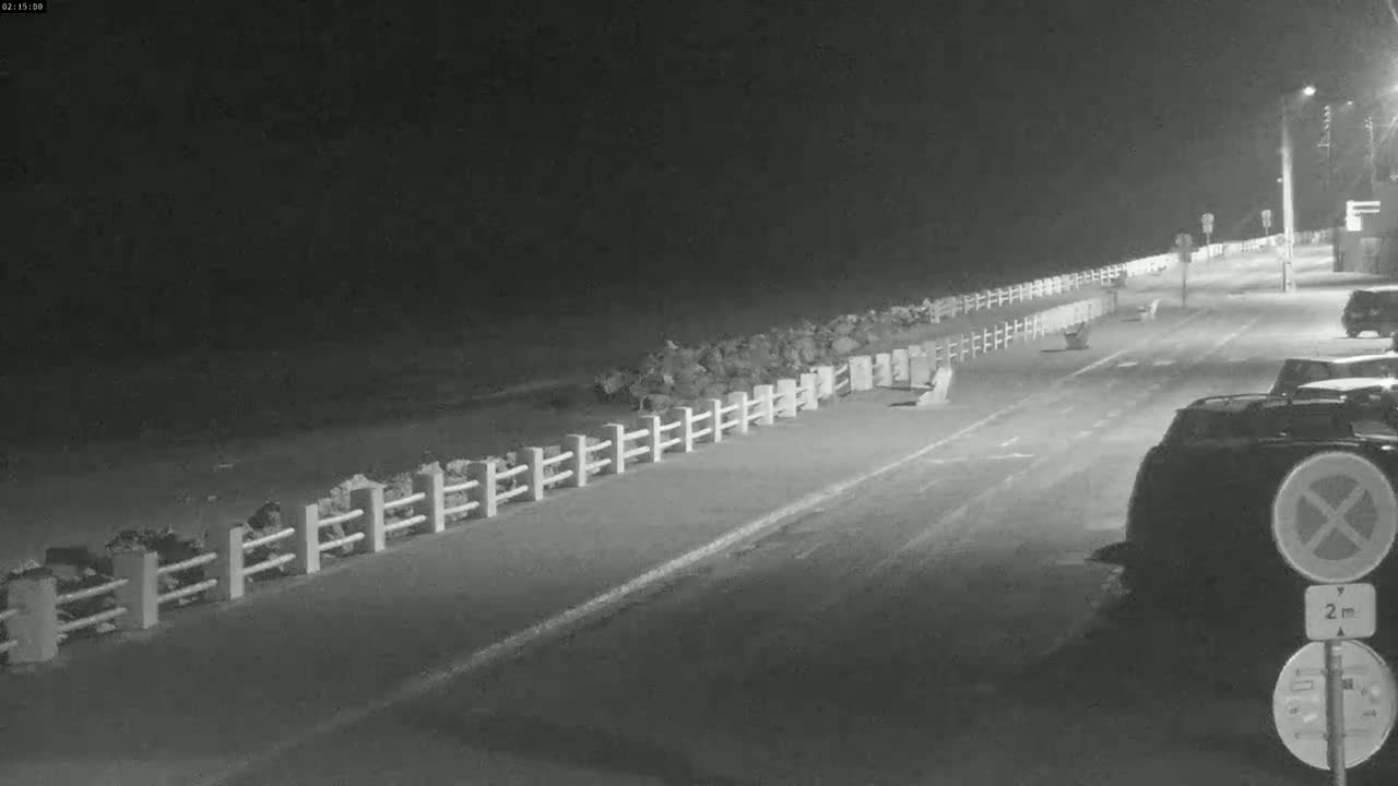A nighttime, low-light view of a road next to a body of water, separated by a low fence and rock barrier, with several parked cars visible.