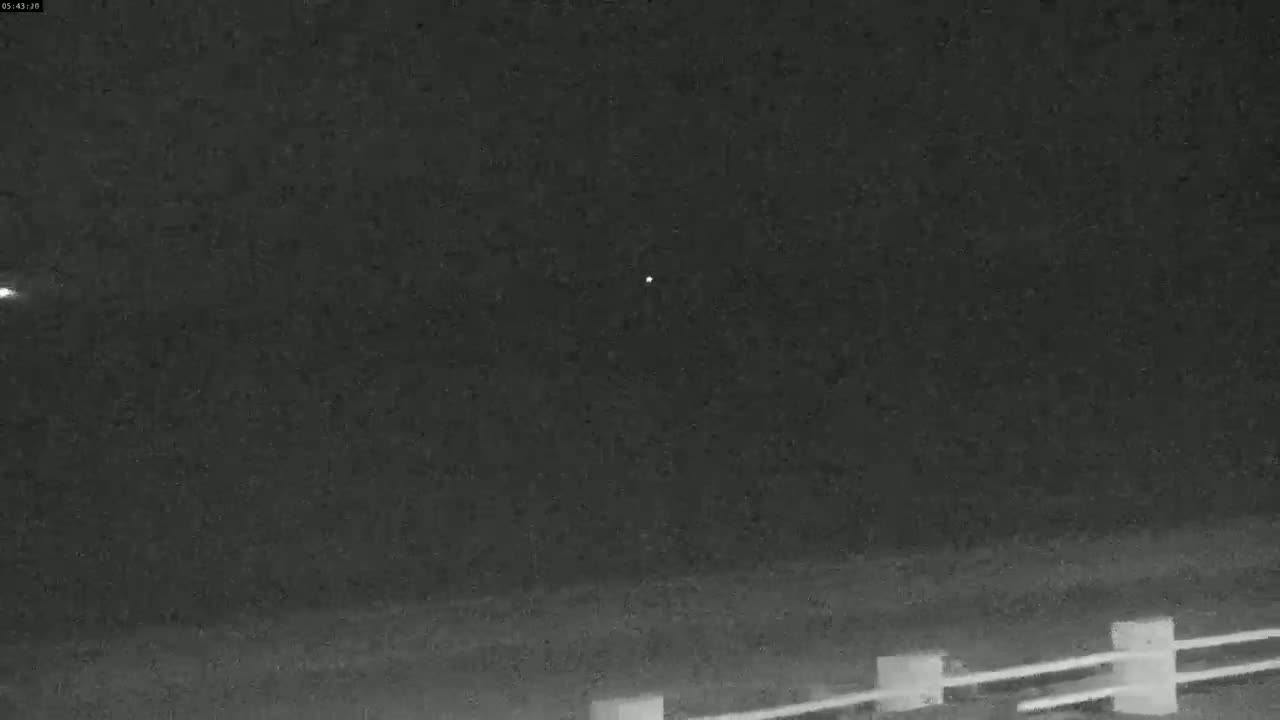 A nighttime, low-light image shows a dark sky with two small, bright lights, one of which appears to have a slight trail, above a low fence.