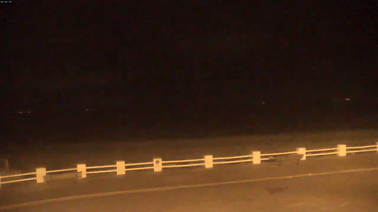 A nighttime view of a road with a low, white fence, extending to a dark horizon under a clear, dark sky.