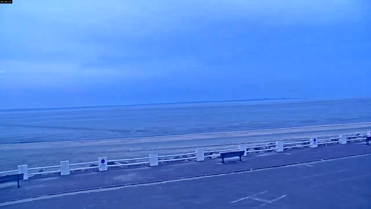 A flat, sandy beach with a low white fence and several park benches is visible under a clear, pale blue sky.