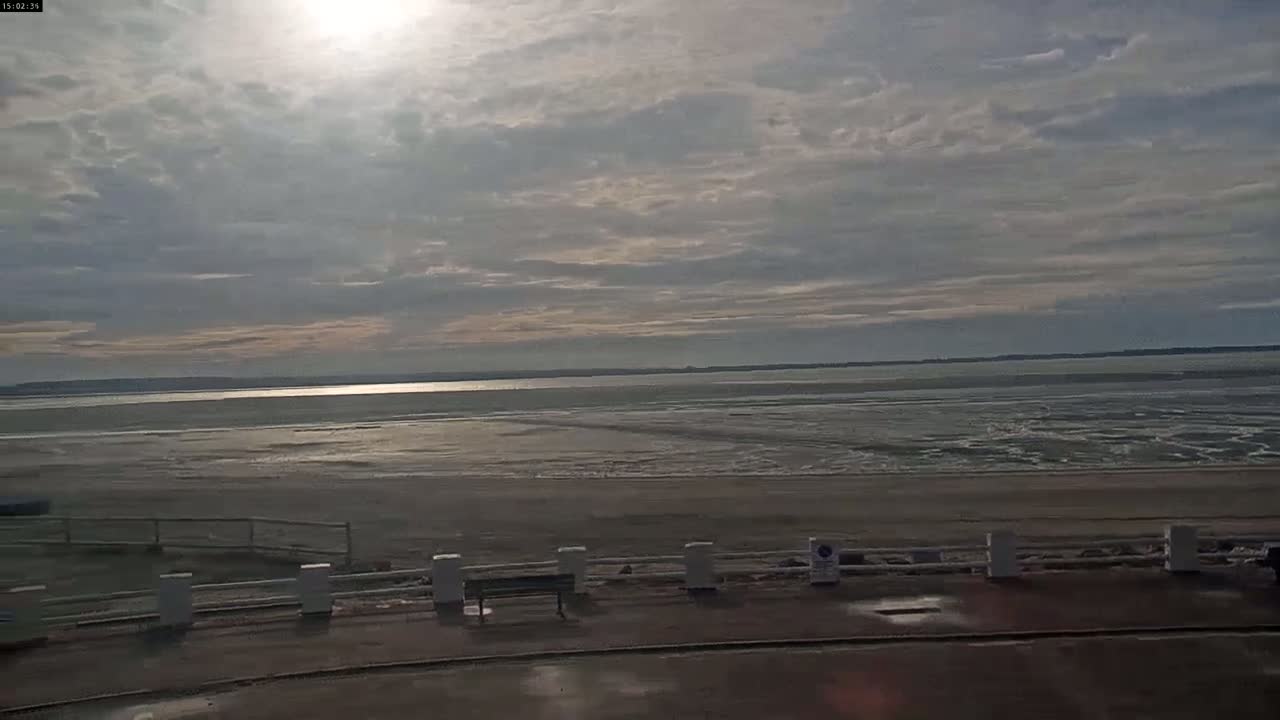 A mostly cloudy day reveals a wide expanse of tidal flats leading to a calm body of water under a bright sun.