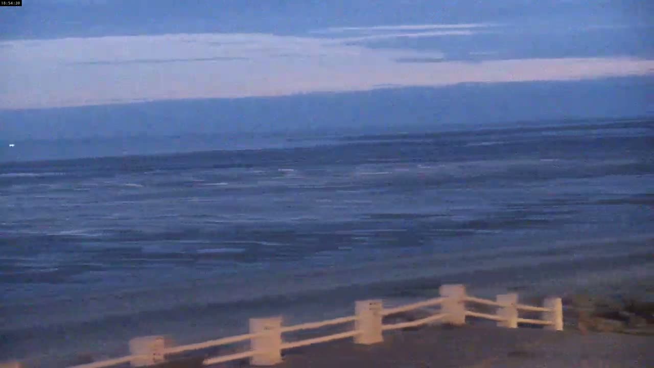 A calm, dark blue ocean stretches to the horizon under a twilight sky, with a low white fence bordering a sandy beach in the foreground.