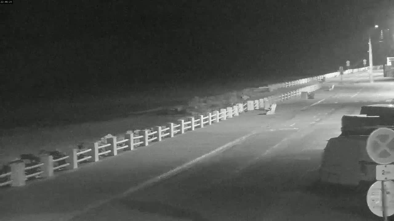 A nighttime, monochrome image shows a paved road next to a beach, separated by a low white fence, with several vehicles parked at the roadside.