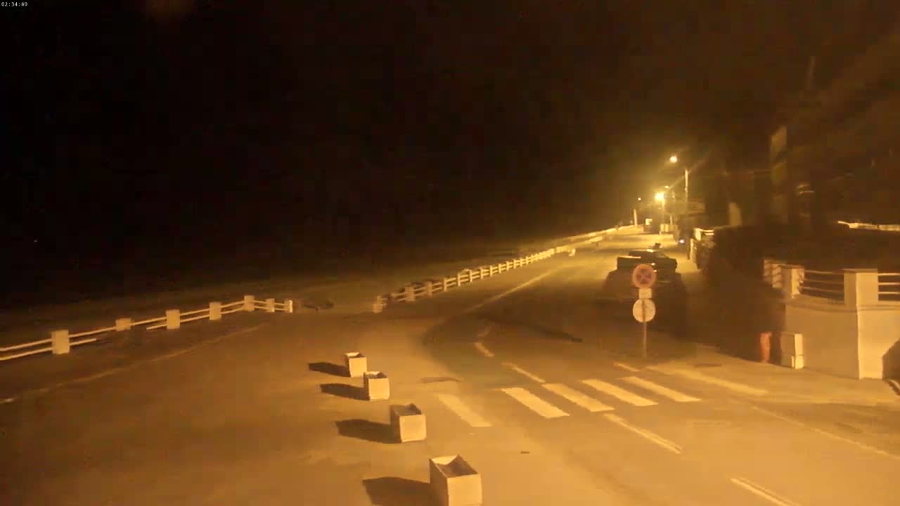 A nighttime view of a paved road next to a beach, bordered by a low white fence, with a few cars parked near buildings under artificial lighting.