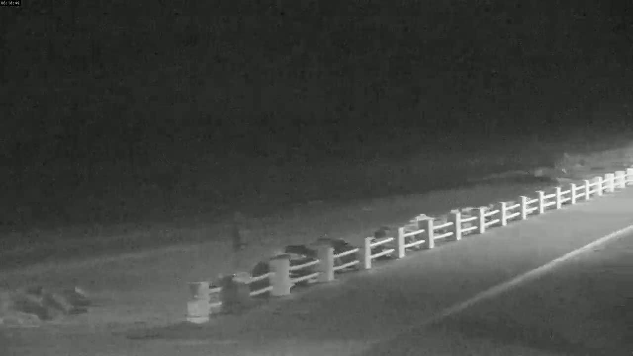 A nighttime, low-light image shows several dark objects, possibly vehicles, parked behind a white fence alongside a road.