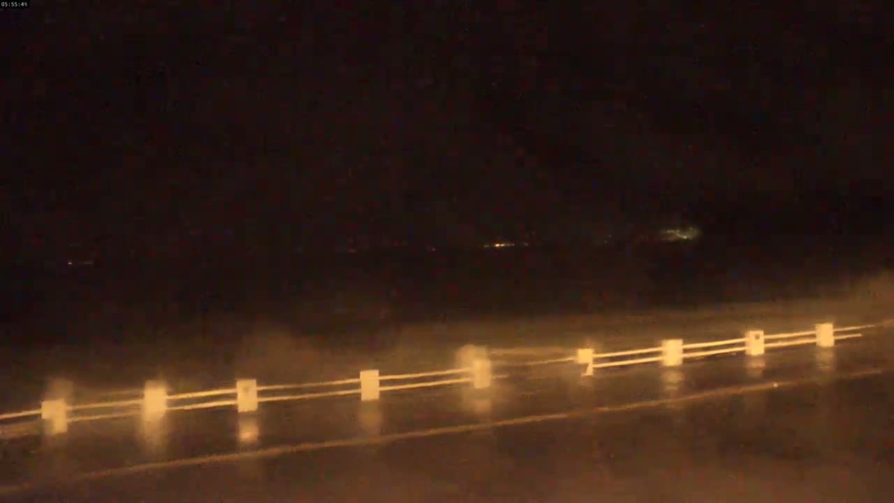 A rain-slicked road at night is visible, showing a low barrier in the foreground and faint lights in the distance.