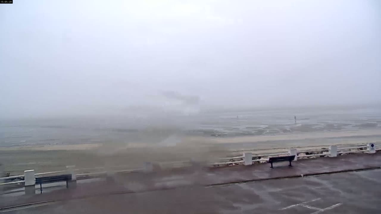 A foggy, overcast day shows a wet, empty seaside promenade with benches and a low white fence separating it from the beach and the water.