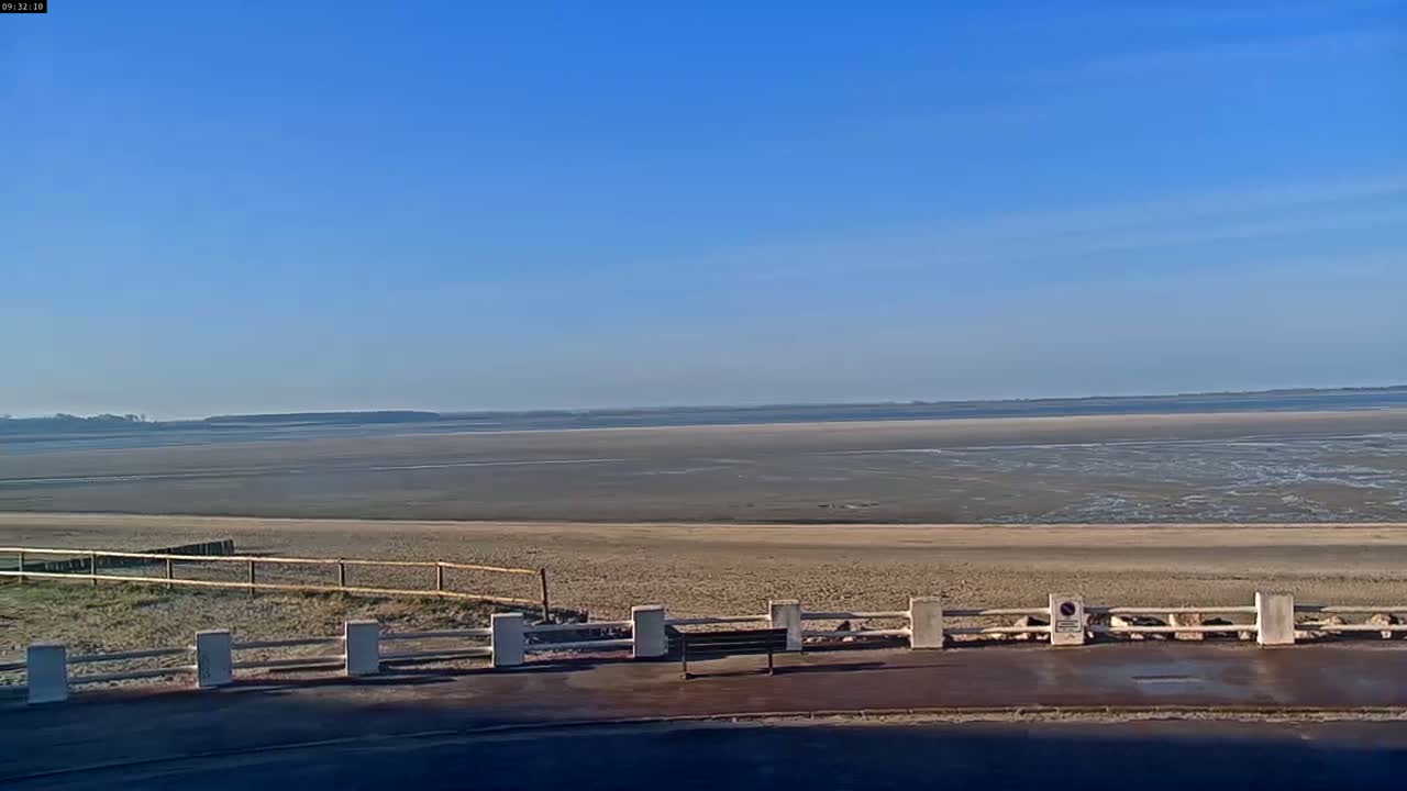 A bench sits behind a low fence overlooking a wide expanse of sandy beach and a calm, flat bay under a clear blue sky.