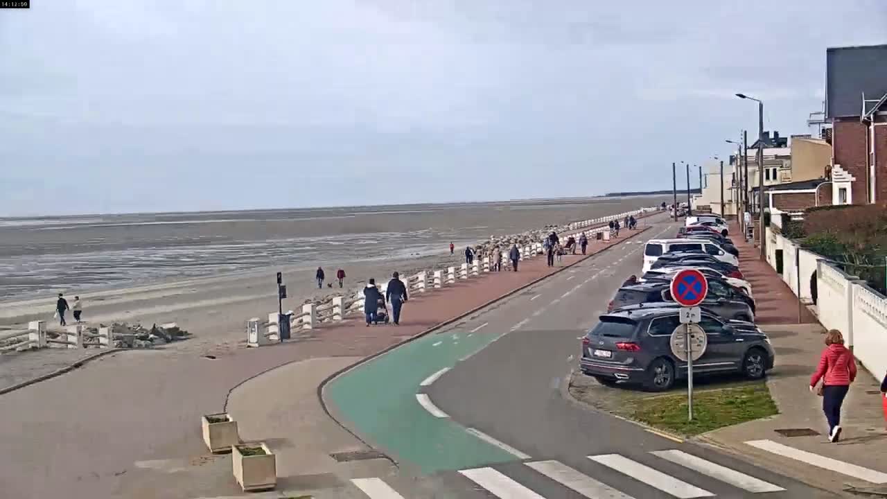 A seaside promenade with pedestrians and parked cars under a cloudy sky, features a wide sandy beach at low tide.