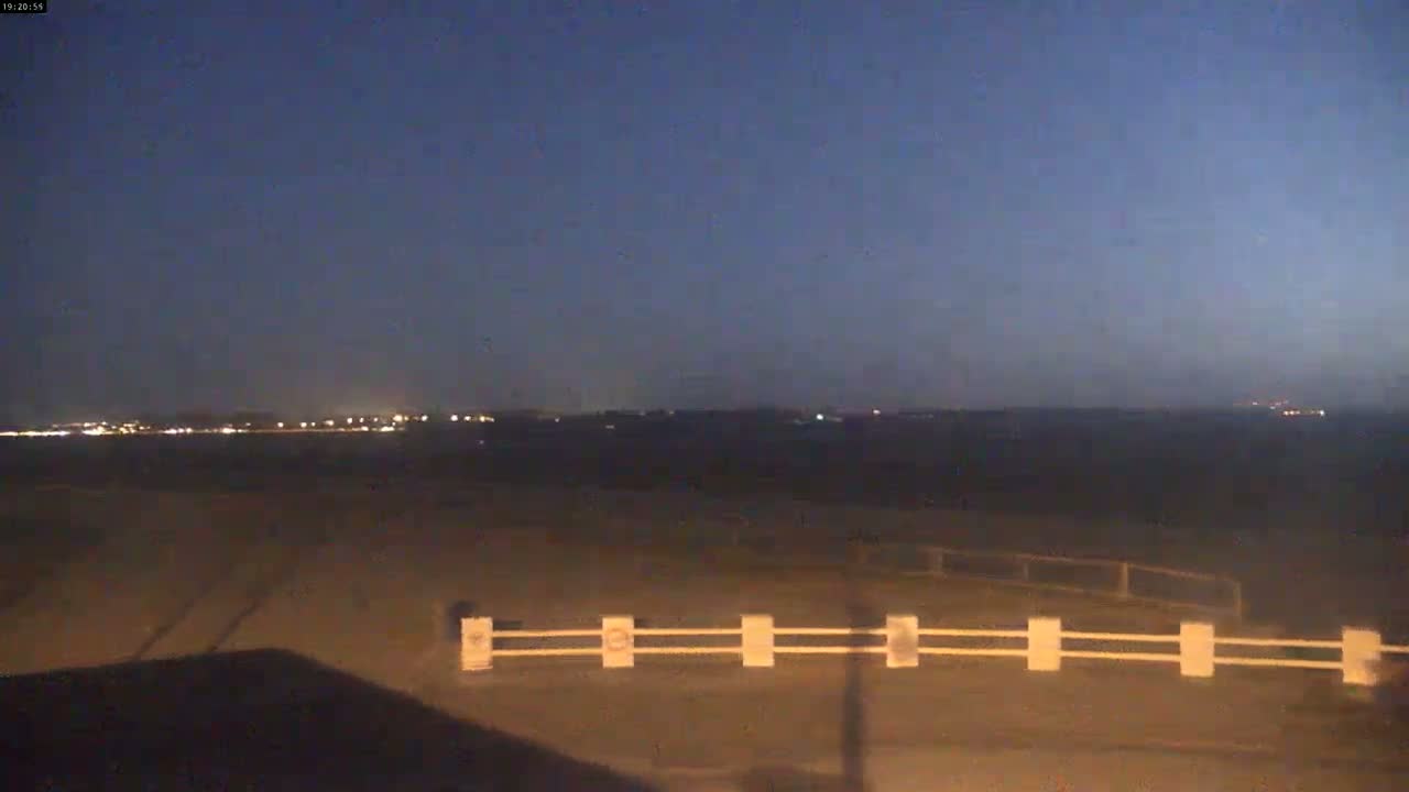 A low-light, twilight view shows a flat, dark landscape with a low white fence in the foreground and distant lights on the horizon.