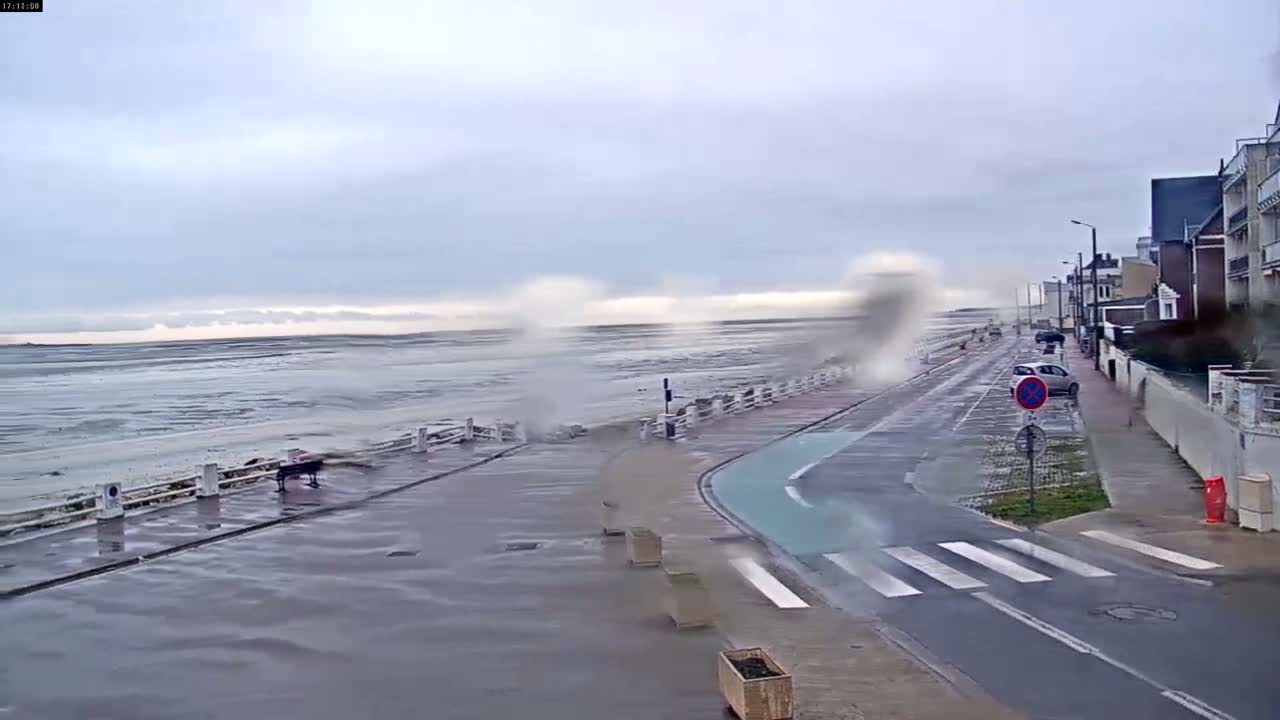 A wet road next to a seawall with crashing waves and some parked cars in the distance, under an overcast sky.