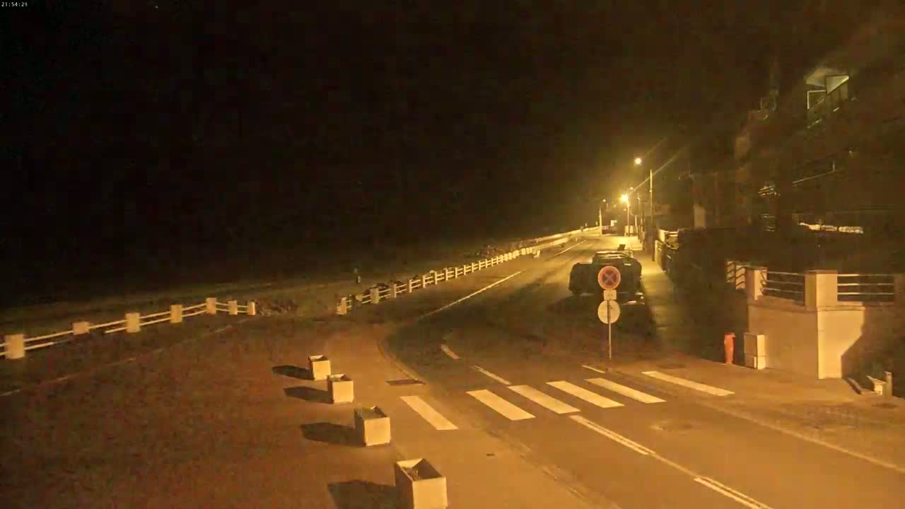 A nighttime view of a mostly empty road next to a beach, lit by streetlights, with a few parked cars and some square planters along the roadside.