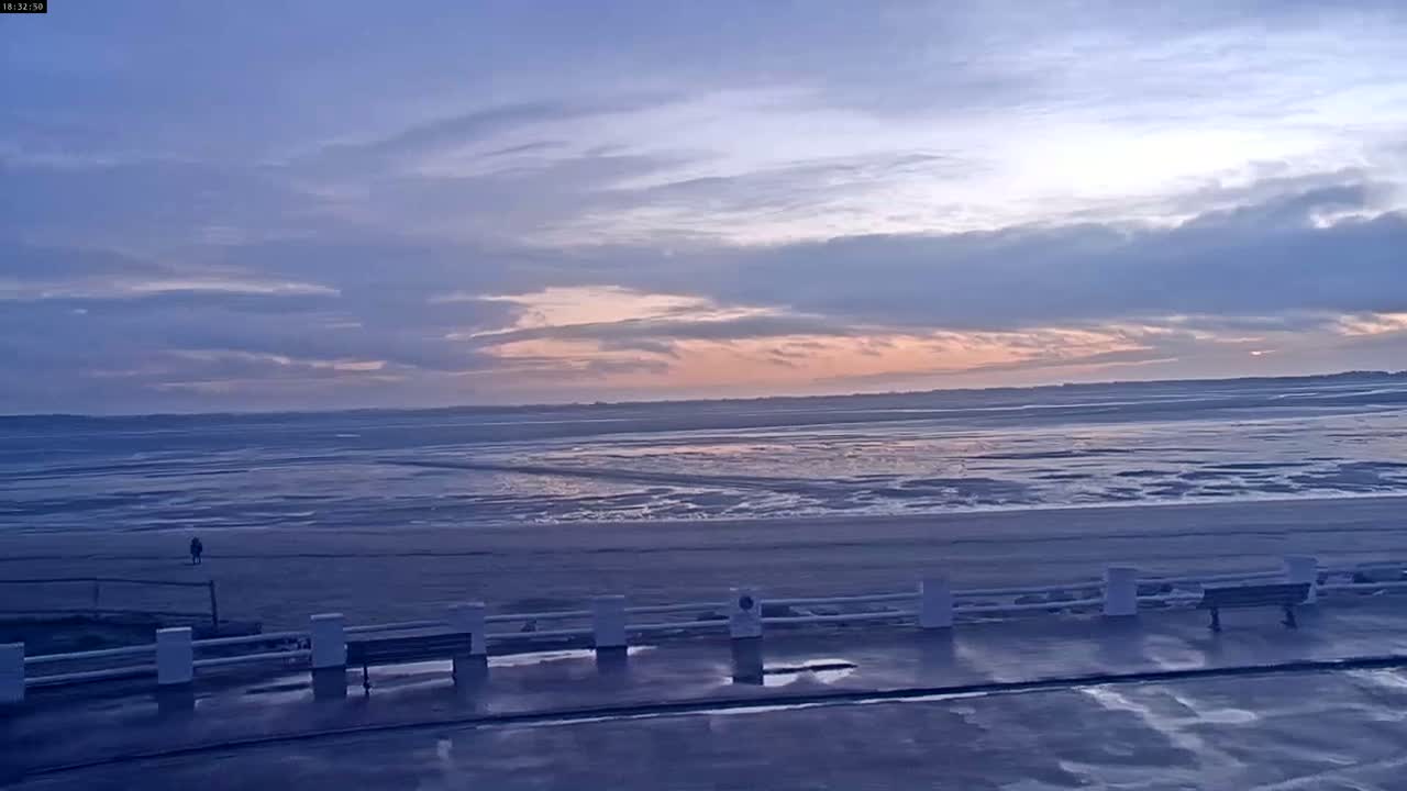 A person stands on a wet beach near a low white fence as the sun sets over a wide expanse of shallow water under a mostly cloudy sky.