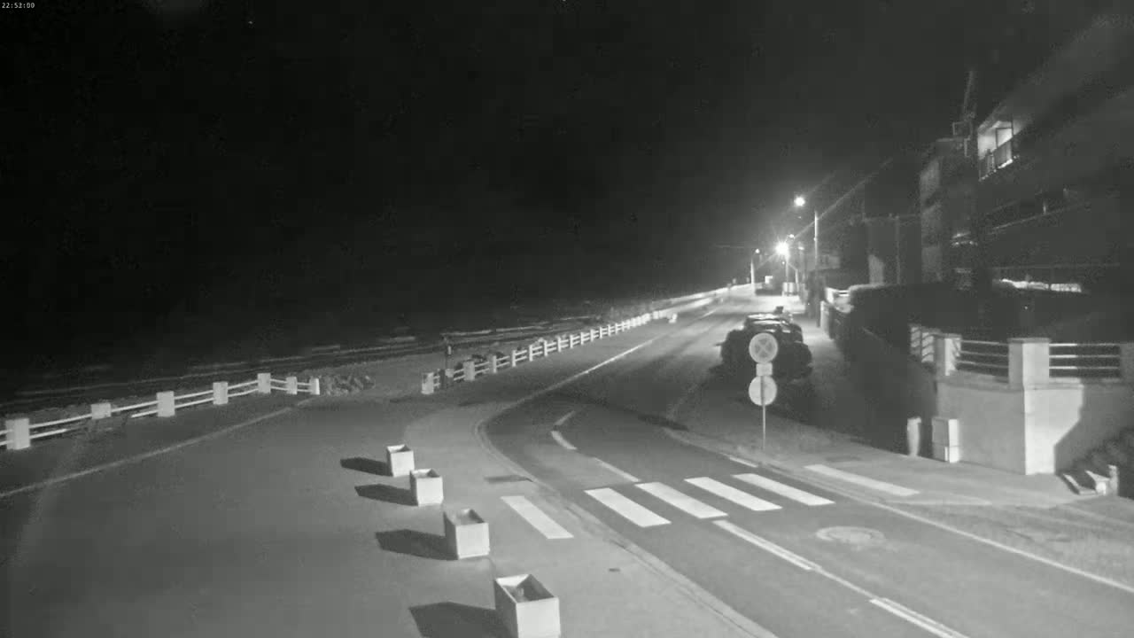 A nighttime, dimly lit street curves near a beach, bordered by a low fence and buildings, with a vehicle parked near the side of the road.
