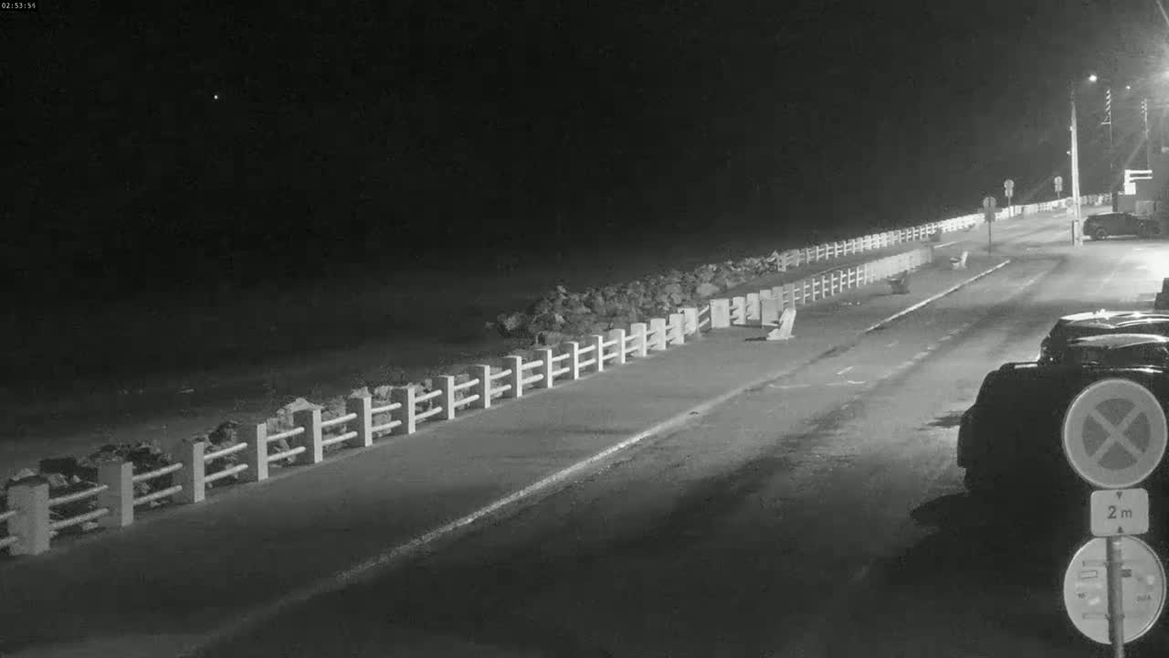A nighttime, low-light view of a paved road next to a low stone wall and fence, with several parked cars visible.